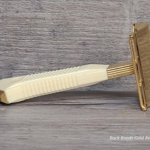 May include: A vintage safety razor with a cream-colored, ribbed handle and gold-colored accents. The razor head and connecting parts are also gold-colored. The image is set against a wooden background. The text "Back Roads Gold Replating" is visible.