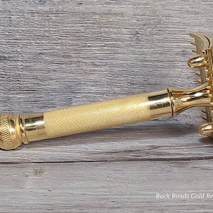 May include: A gold-colored safety razor with a textured handle and a comb-like head. The razor is lying horizontally on a wooden surface. The words "Back Roads Gold Replating" are visible.