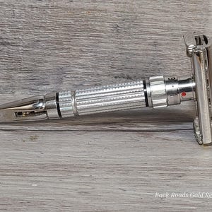 May include: A silver safety razor with a textured handle and adjustable settings. The razor is made of metal and has a classic design, suitable for shaving. The image is set against a wooden background.
