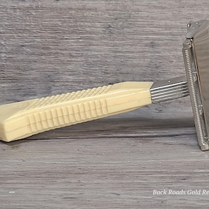 May include: A vintage safety razor with a cream-colored, ribbed handle and a silver-colored head. The razor is designed for shaving and features a classic design. The handle has a rectangular shape with a textured grip.