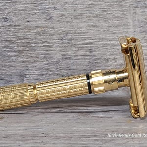 May include: A gold-colored safety razor with a textured handle and adjustable settings. The razor has a classic design and is made of shiny metal. The text "Back Roads Gold Replating" is visible on the bottom right.