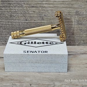 May include: A gold-colored safety razor resting on a white box with the word "Gillette" and "Senator" printed on it. The razor has a textured handle and a classic design. The box has a speckled pattern.