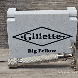 May include: A silver-colored safety razor with a textured handle and a razor head, next to a speckled gray box with the Gillette logo and "Big Fellow" text. The image is on a wooden surface.