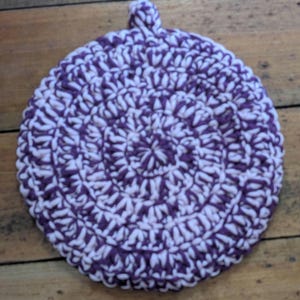 Könnte beinhalten: Ein runder, handgehäkelter Topflappen mit Spiralmuster. Der Topflappen ist hauptsächlich weiß mit einem tiefvioletten Muster. Oben ist eine kleine Schlaufe zum Aufhängen angebracht. Er liegt auf einer Holzoberfläche.