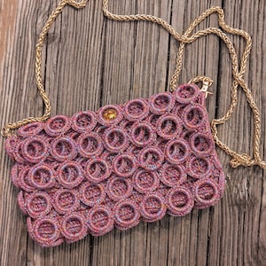 May include: A rectangular handbag crafted from interconnected circular rings in shades of pink, purple, and blue. The bag features a gold-tone metal chain strap and a gold-tone snap closure at the top center, showcasing a textured, woven appearance.