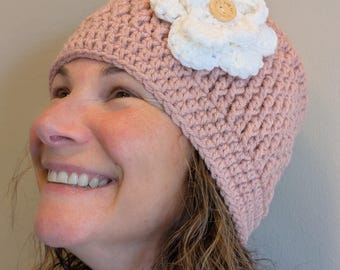 May include: A handmade, light pink crochet hat with a large white flower and a wooden button center. The hat is a beanie style, perfect for cooler weather. The flower adds a decorative touch to the warm, textured hat.
