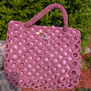 Könnte beinhalten: Eine quadratische Handtasche mit einem altrosa Häkelüberzug in einem sich wiederholenden Kreismuster. Die Tasche hat ein einfarbiges rosa Innenfutter und einen einzelnen passenden Häkelgriff. Eine silberfarbene Blumenbrosche mit zwei hellrosa Steinen ist befestigt.