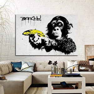 May include: A black and white Banksy print featuring a chimpanzee holding a yellow banana. The word "BANKSY" is stenciled in black above the chimpanzee. The artwork is displayed on a white wall in a living room setting.