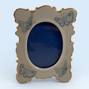 May include: An oval-shaped picture frame with a cream-colored border and gold trim. The frame features four light blue butterfly illustrations. The center of the frame is a deep blue color.