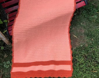 May include: A crocheted throw blanket in shades of peach and orange with a scalloped edge. The blanket is folded and draped over a red wooden bench.
