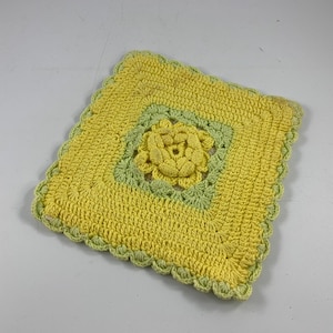 Op de afbeelding: Een vierkante gehaakte onderzetter in geel en groen met een bloemmotief in het midden.