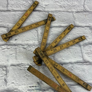 May include: A group of vintage wooden folding rulers with red and black markings. The rulers are laid out on a white brick background.