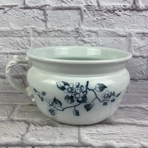 May include: White ceramic chamber pot with a floral design in black and white. The pot has a single handle and a rounded body.