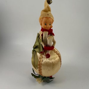 May include: A vintage Christmas ornament featuring a felt elf holding a green and gold package. The elf is wearing a red scarf and a white hat with a gold bell. The ornament is made of a cream-colored fabric with gold glitter accents.
