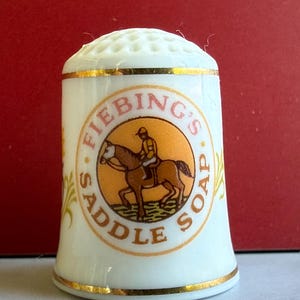 Puede incluir: Un dedal de porcelana blanca con borde dorado y parte superior con hoyuelos. El dedal presenta un diseño circular con el texto "Fiebing's Saddle Soap" alrededor de una imagen de una persona montando a caballo. Acentos dorados decorativos en el lateral.