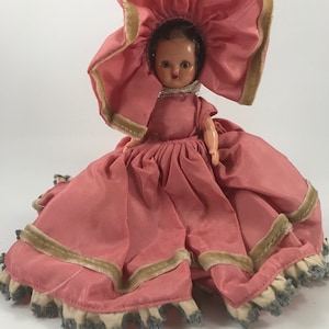 May include: A vintage doll wearing a pink dress with a large pink hat and a white lace collar. The doll has brown hair and painted brown eyes.