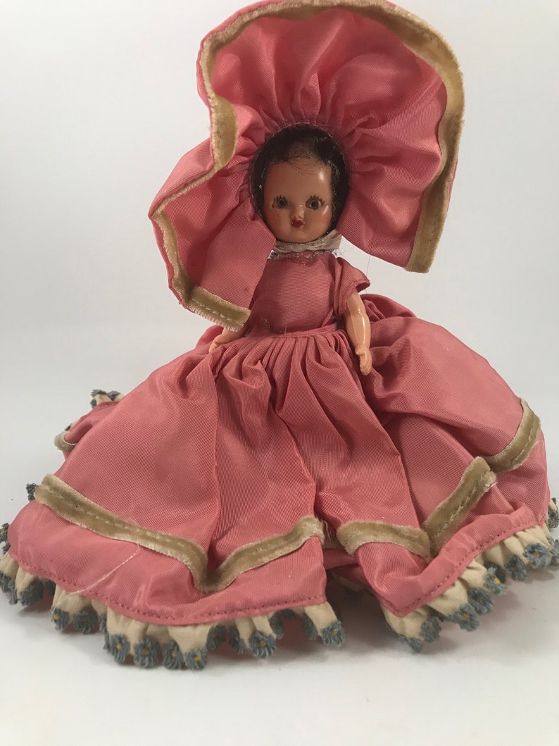 May include: A vintage doll wearing a pink dress with a large pink hat and a white lace collar. The doll has brown hair and painted brown eyes.