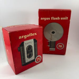 May include: Two red boxes with vintage cameras and flash units. The box on the left has a black and silver camera with the text "argoflex Seventy-five" on it. The box on the right has a flash unit with the text "argus flash unit" on it.