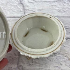 May include: White porcelain trinket box with a gold trim and a scalloped edge. The box is open and the inside is visible. The box has three small feet.