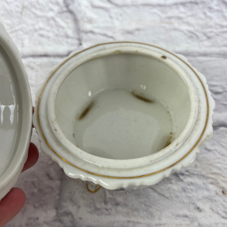 May include: White porcelain trinket box with a gold trim and a scalloped edge. The box is open and the inside is visible. The box has three small feet.