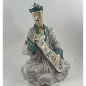 Puede incluir: Figura de cerámica de una figura asiática sentada con una túnica gris claro con detalles turquesa. La figura sostiene un pergamino con caracteres asiáticos. La figura tiene una expresión serena y lleva un sombrero con una banda turquesa.