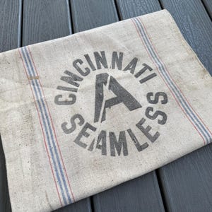 May include: A vintage, off-white fabric sack with the words "CINCINNATI SEAMLESS" printed in gray. The sack has red and blue stripes along the edges. The fabric appears worn, giving it a rustic aesthetic.