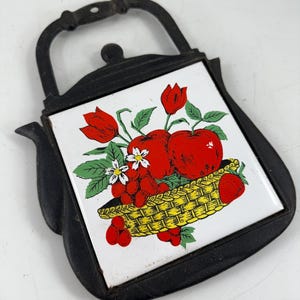 May include: Black cast iron trivet in the shape of a teapot. The center tile features a colorful illustration of red apples, strawberries, and flowers in a yellow basket. The trivet has a handle for easy carrying.