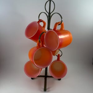 Könnte beinhalten: Ein Set aus sechs orangefarbenen Tassen mit rosa Innenräumen, die an einem dunklen Metall-Tassenbaum hängen. Die Tassen haben eine glänzende Oberfläche und sind kreisförmig angeordnet. Der Tassenbaum hat ein dekoratives, wirbelndes Design.