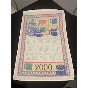 Op de afbeelding: Crèmekleurige keukendoek met een kalenderontwerp voor het jaar 2000. De doek toont illustraties van koffiepotten en kopjes in blauw, groen en rood, met een rand van blauwe en roze ruitpatronen.