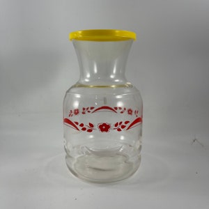 Puede incluir: Una jarra de vidrio transparente con tapa amarilla y diseño floral rojo. La jarra tiene una base ancha y un cuello estrecho.