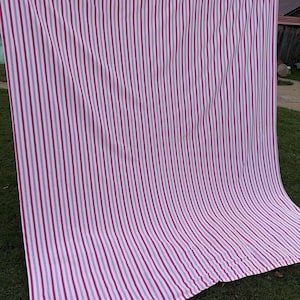 May include: A large, striped curtain or fabric panel, mainly white with vertical stripes in varying shades of pink and magenta. The top edge is a solid magenta colour. The fabric is hanging outdoors, possibly drying on a washing line.