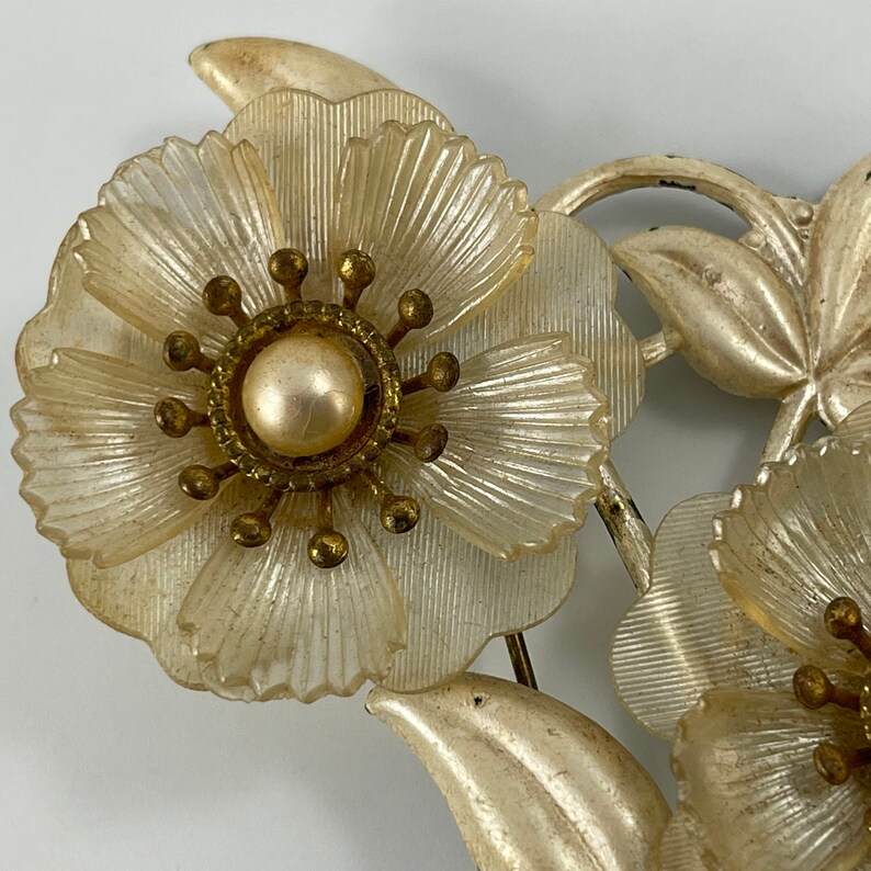 May include: A vintage brooch featuring three white plastic flowers with a pearl centre and gold accents. The flowers are arranged in a cluster with leaves and stems.