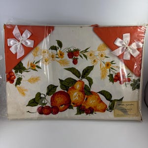 Puede incluir: Un juego de mantel vintage con un diseño de frutas, que incluye peras, manzanas y bayas. El mantel blanco tiene un borde floral y está empaquetado con dos esquinas naranjas adornadas con lazos blancos. El juego está sellado en plástico.