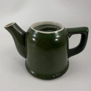 May include: A dark green ceramic teapot with a matching lid and handle. The teapot has a spout and a white rim around the top. The handle is a curved, open design. The teapot is a classic shape and design.