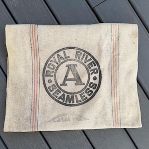 May include: A vintage, off-white canvas bag with red and blue stripes. The bag has a black circular logo that reads "Royal River Seamless" with a large letter "A" in the center.