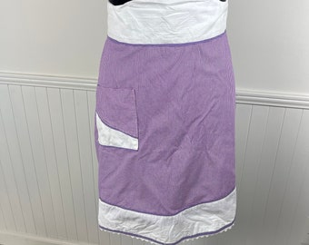 May include: A purple and white gingham apron with a white pocket and white trim. The apron has a scalloped edge.