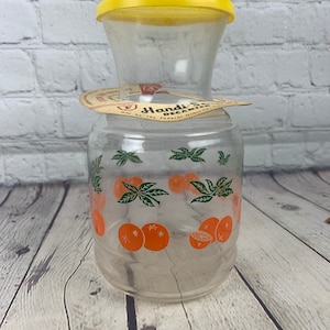 Puede incluir: Una jarra de vidrio transparente con tapa amarilla y diseño naranja y verde. La jarra tiene una etiqueta que dice "Handi-Pour Decanter".
