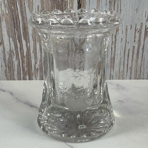 Può includere: Un vaso di vetro trasparente per boccioli con un bordo testurizzato a perline e un motivo floreale. Il vaso è alto circa 10 cm.