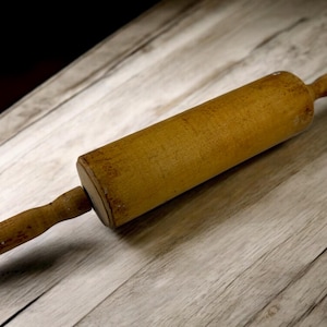 May include: A wooden rolling pin with a cylindrical body and two handles. The rolling pin is light brown and appears to be well-used, with visible wear and tear. It rests on a light-colored wooden surface, suggesting a kitchen or baking theme.