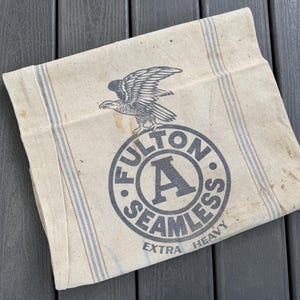 May include: Vintage beige fabric bag with blue stripes and a large circular emblem. The emblem features an eagle above the words "FULTON SEAMLESS EXTRA HEAVY" in a dark gray color. The bag has a worn, aged appearance.