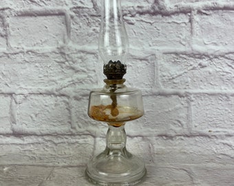 May include: A clear glass oil lamp with a metal top and a round base. The lamp is sitting on a white brick surface.
