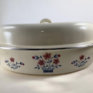 May include: Cream-colored oval roaster with a matching lid. The roaster features a floral design in red, blue, and white. It has two side handles and a loop handle on the lid. The roaster is made of metal.