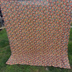 May include: A large rectangular fabric piece featuring a dark brown background with a repeating floral pattern of small orange, red, and light blue flowers connected by thin green vines, displayed hanging vertically against an outdoor backdrop.