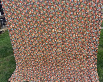 Op de afbeelding: Een groot rechthoekig stuk stof met een donkerbruine achtergrond en een herhalend bloemenpatroon van kleine oranje, rode en lichtblauwe bloemen verbonden door dunne groene ranken, verticaal opgehangen in een buitenomgeving.