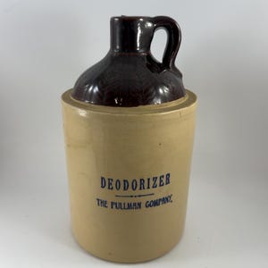 Puede incluir: Una jarra de gres vintage con una parte superior y un asa de color marrón oscuro y texturizado, y un cuerpo de color beige. La jarra presenta el texto "DEODORIZER THE PULLMAN COMPANY" en letras azules, lo que sugiere su propósito original.
