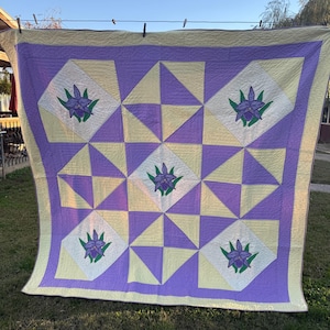 May include: A square quilt featuring a geometric pattern of light yellow and lavender triangles. Four white diamond-shaped patches are centered in the quadrants, each displaying an embroidered purple orchid with green leaves.