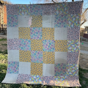 May include: A vintage patchwork quilt with a variety of patterned squares in shades of yellow, purple, blue, and gray. The quilt is hanging outside on a clothesline, showcasing its handmade construction and design. The quilt's dimensions are not visible.