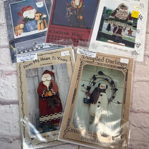 May include: Several Christmas-themed craft patterns in plastic sleeves. The patterns feature Santa Claus, snowmen, and other holiday designs. Some patterns have text like "Santa & Rudy", "Cotton Way", and "From My Heart To Yours".