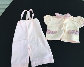 Puede incluir: Un conjunto de bebé rosa con una camisa de manga corta abotonada y un mono a juego. La camisa tiene un cuello y ribetes en los bolsillos a cuadros rosa y blanco. El mono tiene tirantes ajustables.