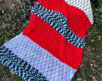 May include: A crocheted blanket with stripes of red, white, and green yarn. The blanket is folded and draped over a wooden bench.